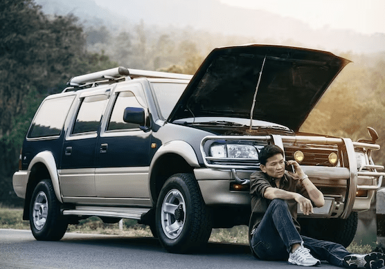 A woman sitting on the side of a road next to an open car hood.